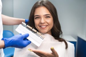 Woman with brown hair in dental chair gesturing to veneer shade guide