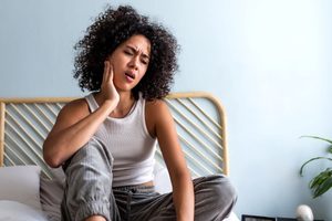 Woman sitting on bed touching sore jaw with hand